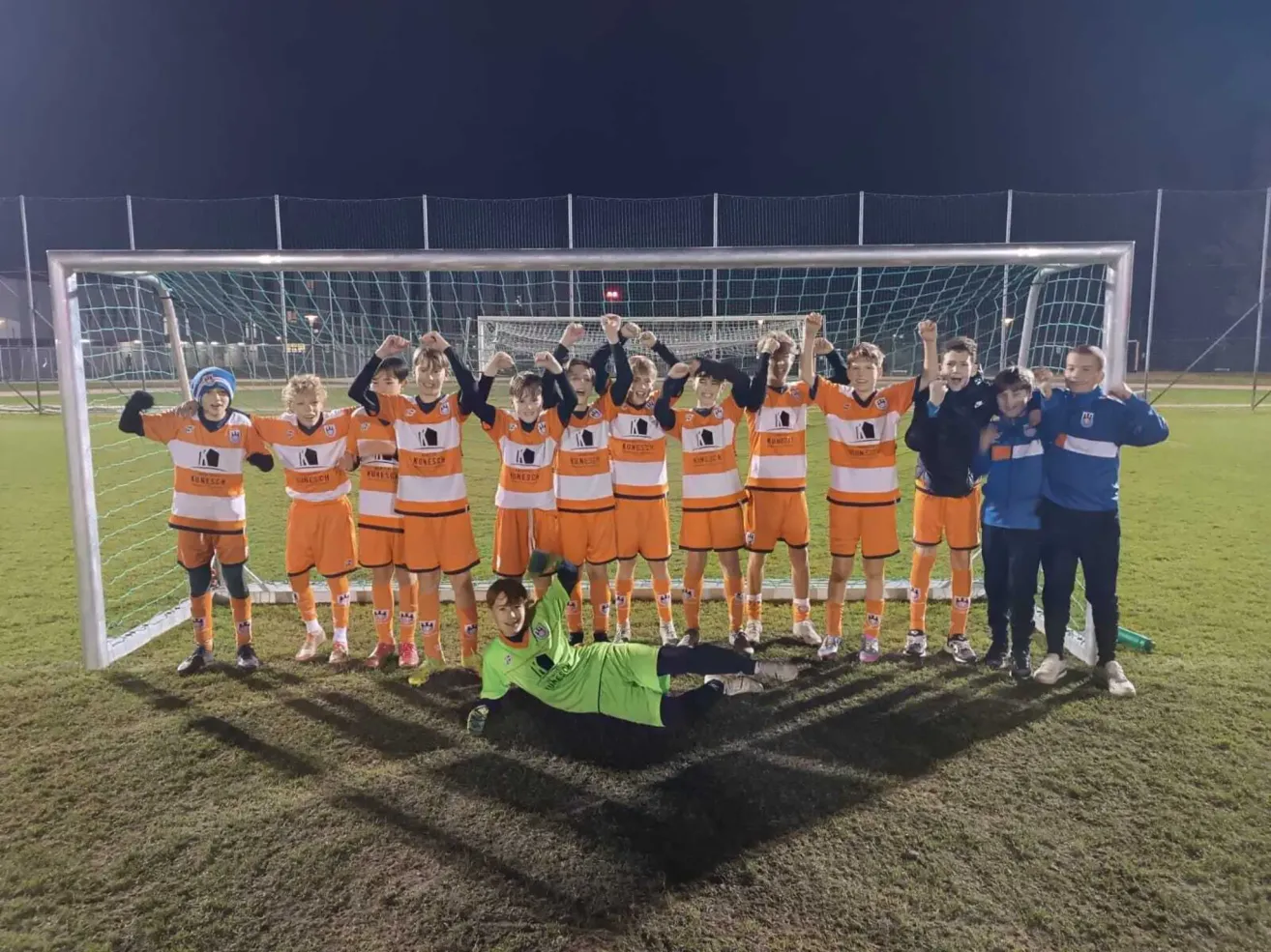 A soccer team is posing for a photo on a field. The players are wearing orange uniforms. The goalkeeper is lying on the ground in front of the goal.