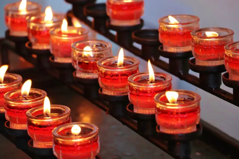Eine Reihe roter Kerzen in klaren Glasbechern, die in einem schwach beleuchteten Raum brennen. Jede Kerze ist in einem schwarzen Metallhalter eingeschlossen und bildet eine Reihe leuchtender Votivkerzen.