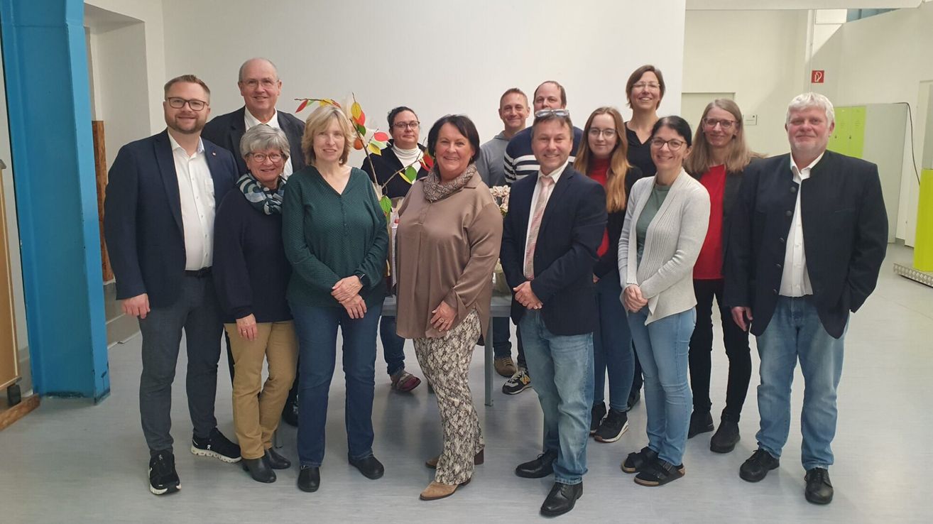A group of people stands together, smiling, and posing for a photo in a white-walled room. A table with leaves is behind them.