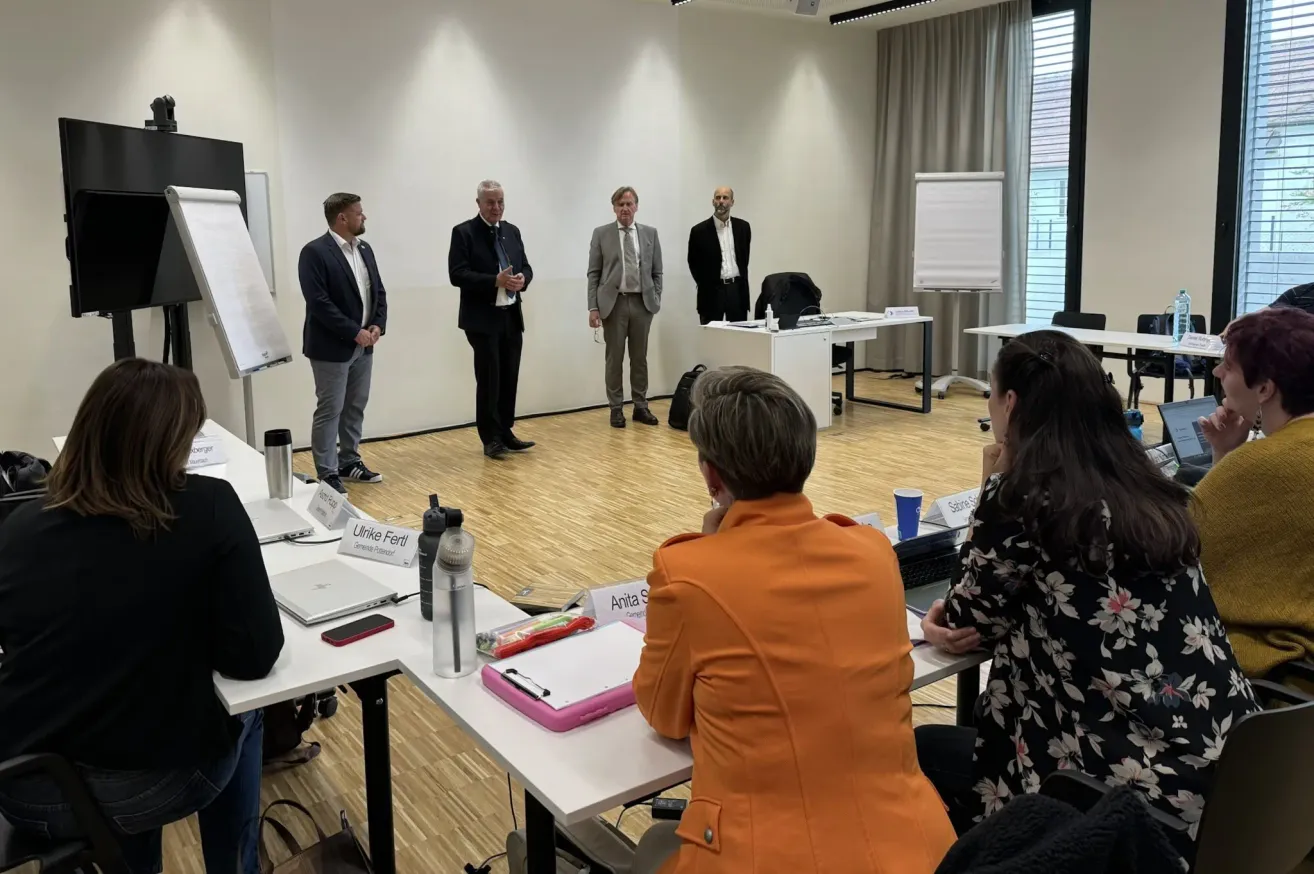 A group of people in formal attire are standing and talking in a conference room. A woman in an orange blazer sits at a table.