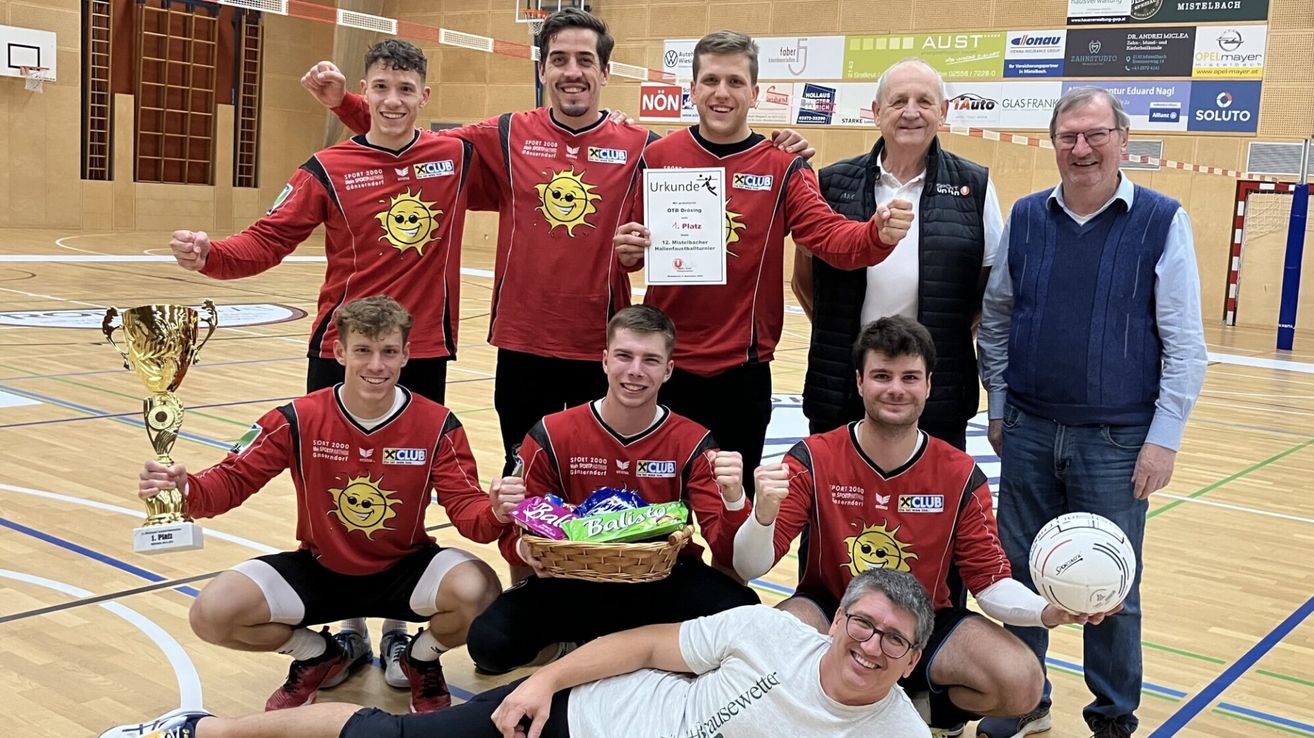 A group of men wearing red jerseys with a yellow sun design are posing for a photo. One man holds a trophy, while another holds a basket filled with candy. They are on a basketball court with a wooden floor and walls with advertisement posters.