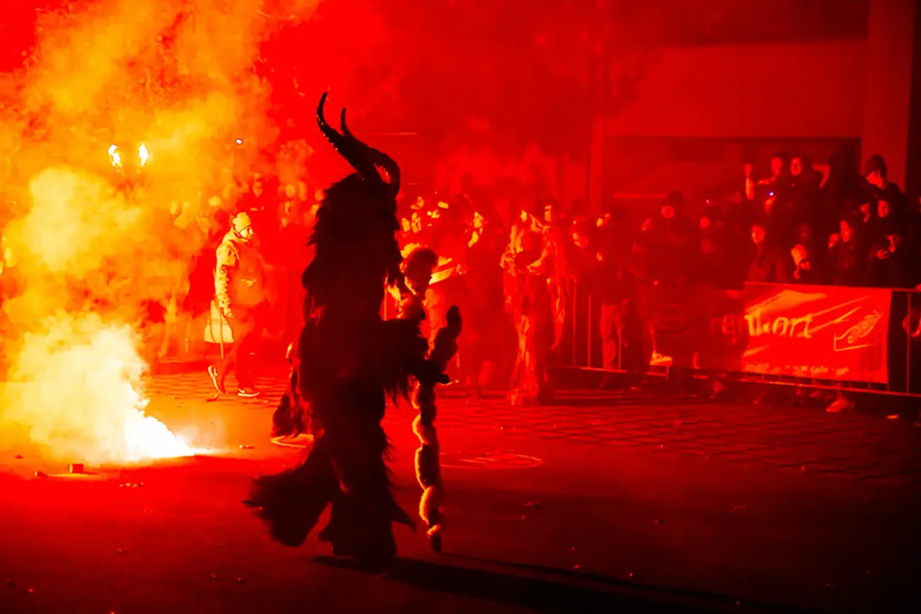 A person in a horned costume stands amidst a crowd, flames in the background, during a lively event.