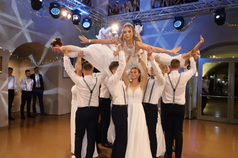 A group of dancers lift a bride on a stage. The bride is wearing a crown and a necklace. The dancers are dressed in white shirts and black suspenders. Spotlights and decorative lights are in the background.
