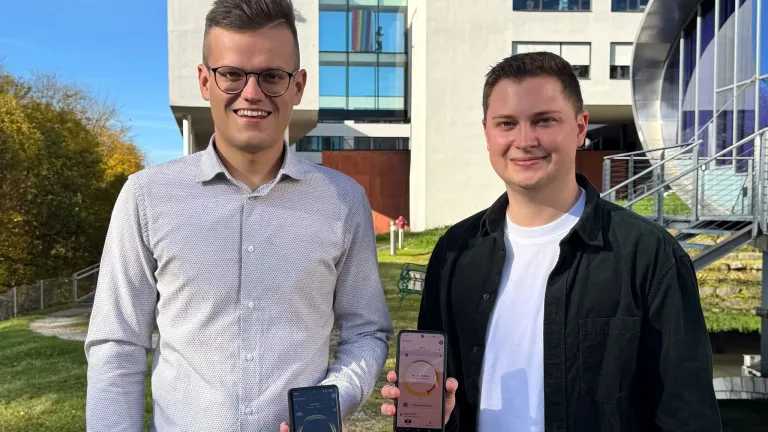 Two men smiling, standing in front of a modern building, each holding a smartphone. The man on the left wears glasses and a light-colored shirt, while the man on the right wears a dark jacket and a white shirt.