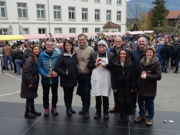 A group of people poses for a photo in front of a large crowd, with a building and mountains in the background.