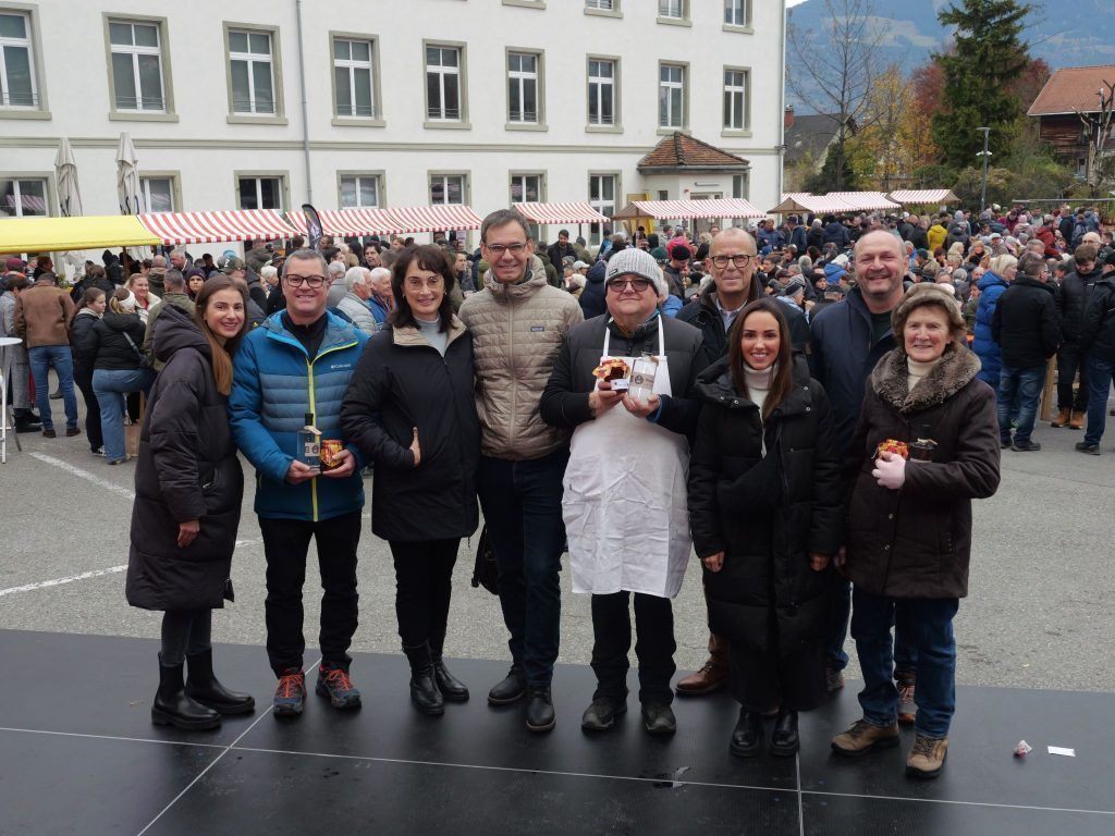 A group of people poses for a photo in front of a large crowd, with a building and mountains in the background.