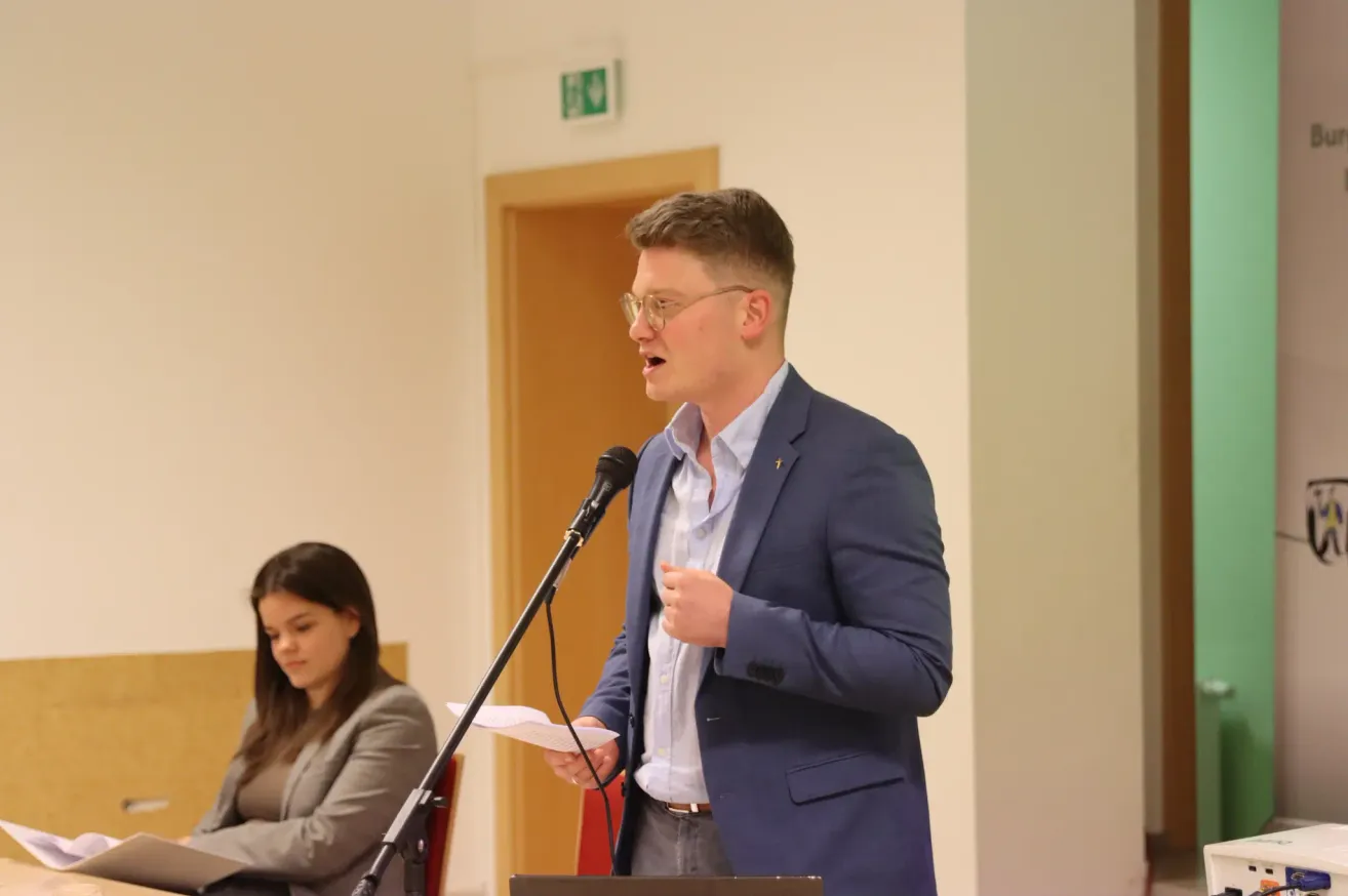 A man speaks into a microphone at a meeting. He holds a paper in his hand. A woman sits behind him. The room has a white wall and a brown door.