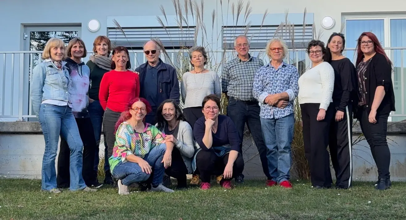 Eine Gruppe Erwachsener steht vor einem Gebäude und posiert für ein Foto. Die Personen lächeln und einige stehen, während andere auf dem Gras sitzen.