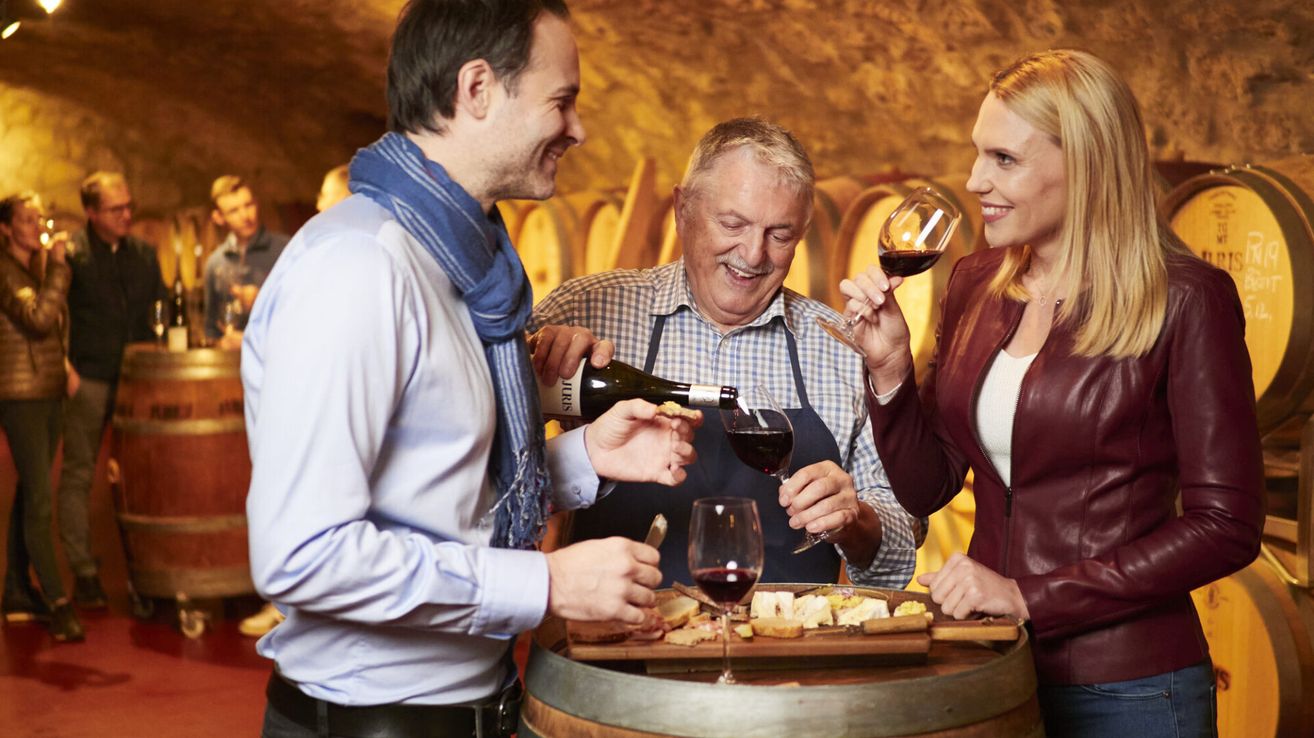 Three people are standing around a barrel filled with cheese and wine. A man is pouring wine into glasses for the others.