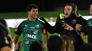 Two soccer players celebrate on the field at night. One wears a green jersey with white letters and the other a black jacket.