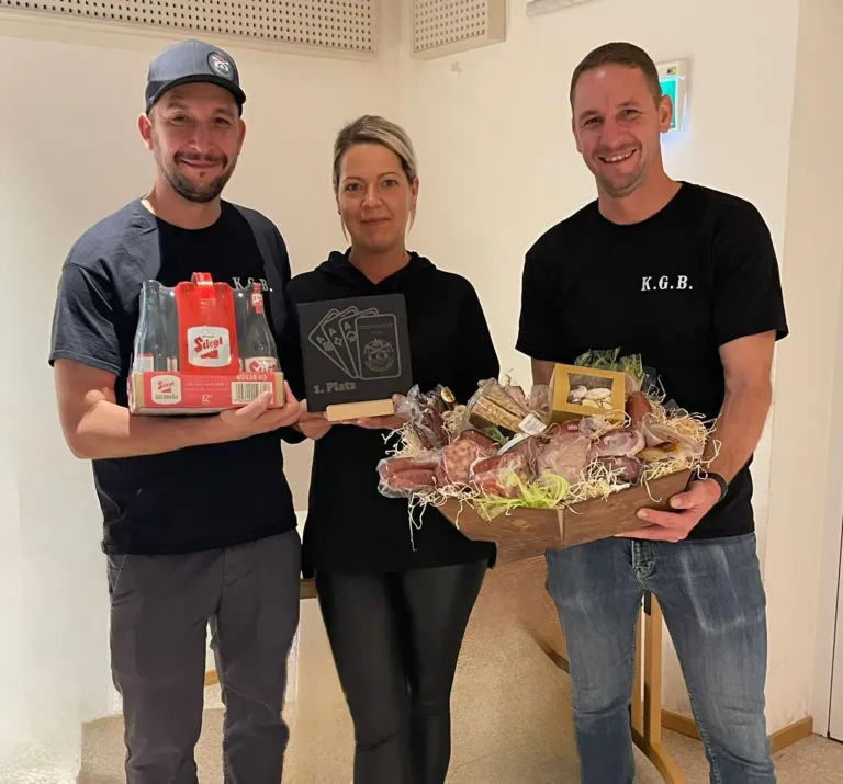 Drei Personen stehen in einem Raum. Der Mann auf der linken Seite hält eine Geschenkbox mit dem Wort 'Stiegl'. Die Person in der Mitte hält ein Buch mit dem Titel 'L. Platz'. Der Mann auf der rechten Seite hält einen Korb mit verschiedenen Gegenständen. Sie alle lächeln für die Kamera.