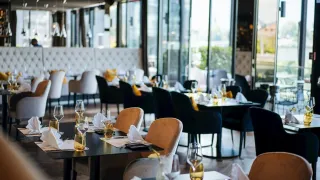 An empty restaurant with large windows and modern furniture. Each table is set with glasses, plates, and napkins. The room is well-lit with natural light.