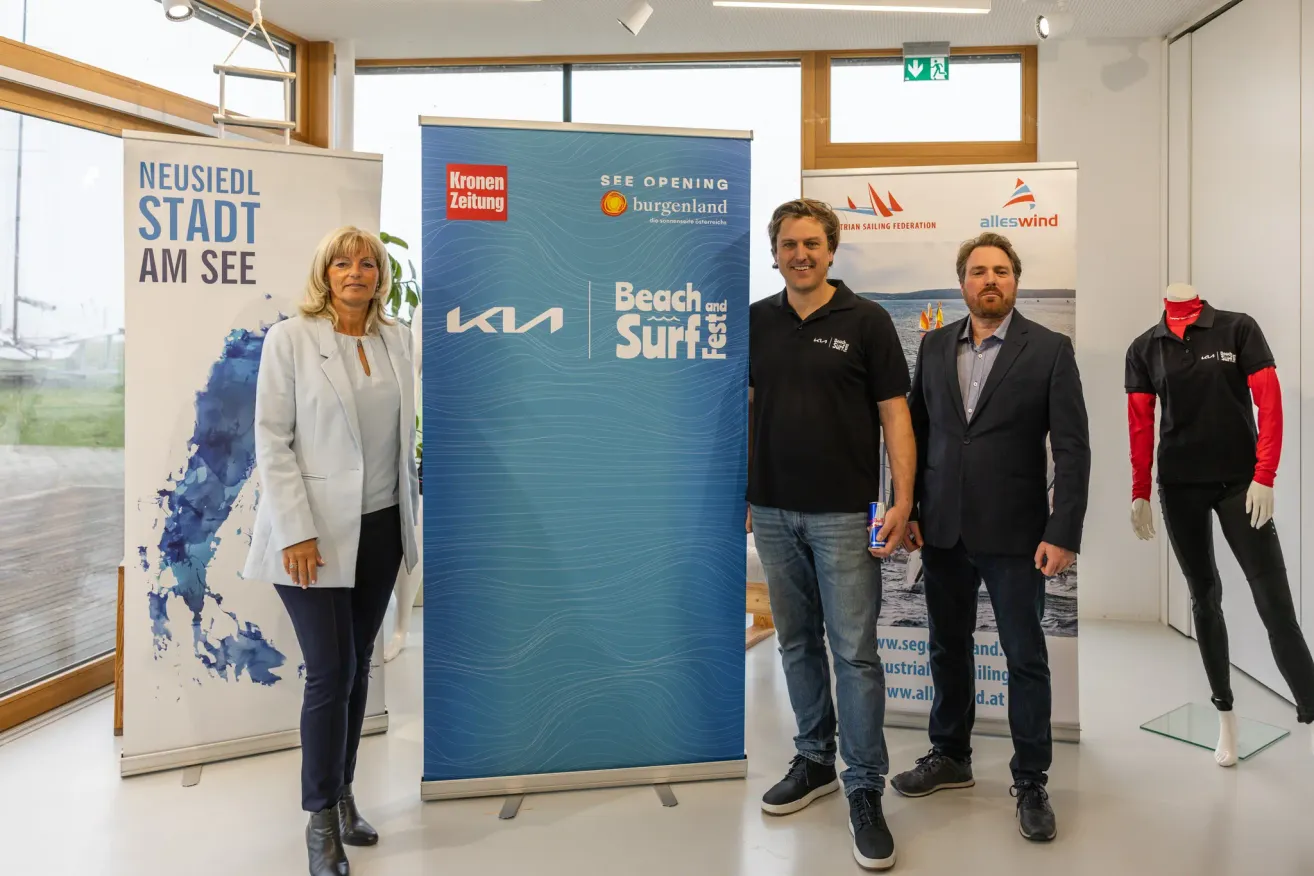 Three people stand in front of a promotional banner, two men and a woman, all smiling for the camera. The banner features a blue wave and the text 'SEE OPENING'.
