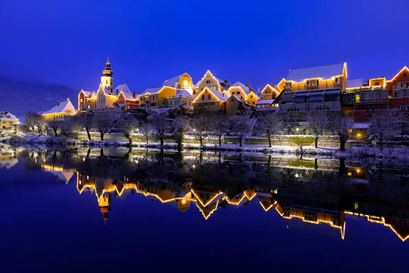A nighttime view of a European town with illuminated buildings, reflecting on the water surface. Snow-covered rooftops and trees enhance the winter ambiance.