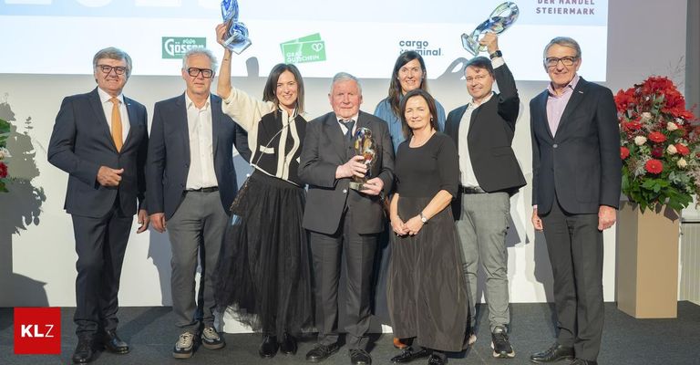 A group of people holding awards are posing for a photo on stage. The man in the middle is holding a golden trophy. Behind them is a white wall with some logos and texts.