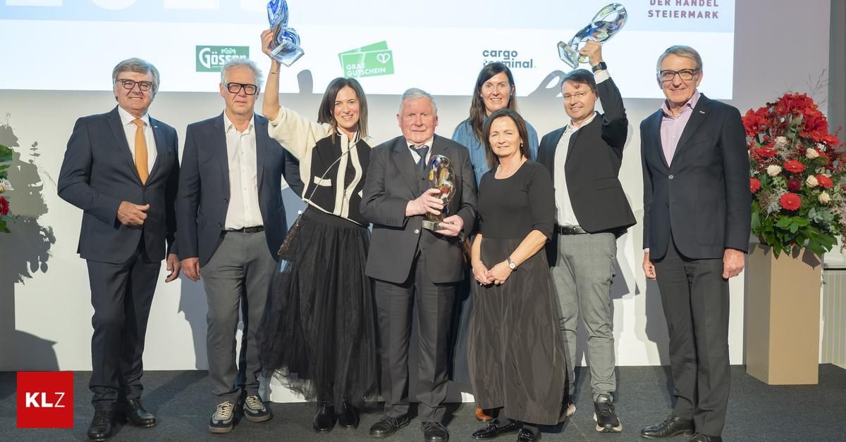 A group of people holding awards are posing for a photo on stage. The man in the middle is holding a golden trophy. Behind them is a white wall with some logos and texts.