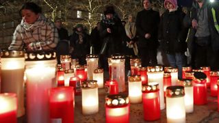 A night scene with people standing around lit candles on a cold evening. Trees and buildings are visible in the background.
