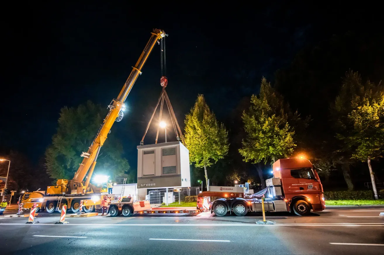 A crane lifts a large white container at night, with a red truck on the right, illuminated by streetlights and headlights. Workers are nearby, and trees and a building are in the background.