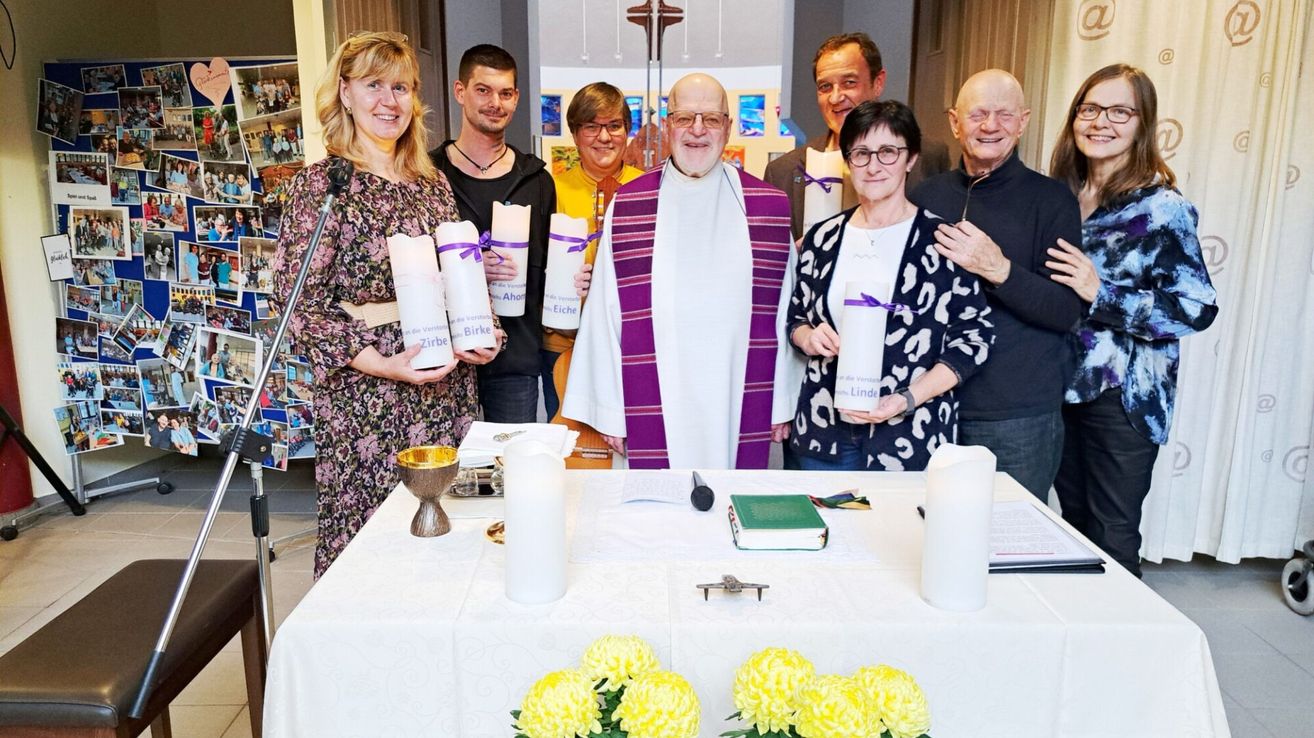 A group of people dressed in religious attire hold candles in a church. A priest stands in the center wearing a white and purple robe. A table with a book and chalice is arranged behind them.