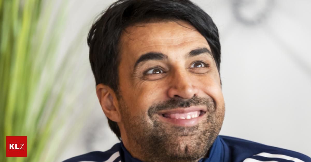 A man with a beard and mustache smiles broadly, his teeth visible, wearing a blue and white jacket. He has dark hair and brown eyes, with a light-colored background behind him.