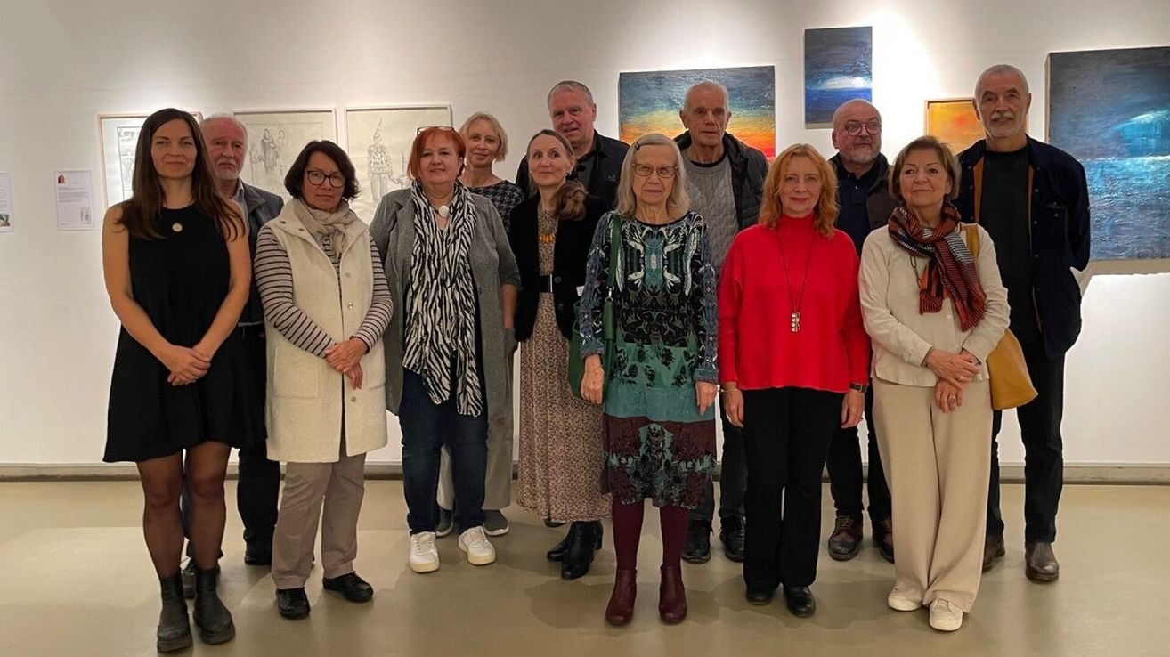 A group of adults stands in front of a white wall with several paintings in an art gallery. They are wearing various clothes and shoes.