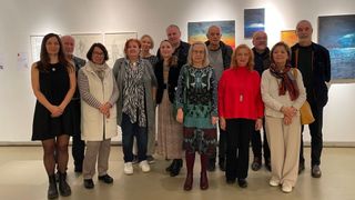 A group of adults stands in front of a white wall with several paintings in an art gallery. They are wearing various clothes and shoes.