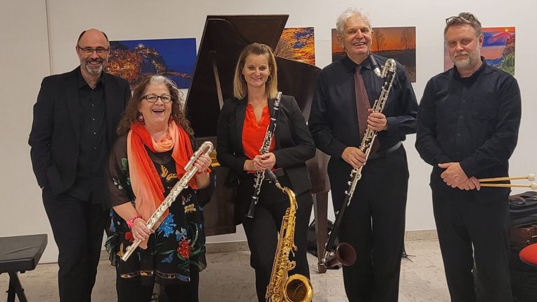 Three musicians stand next to a piano, smiling for a photo. A woman holds a flute, another a saxophone, and a man a clarinet. They are in a room with a gallery-like background.