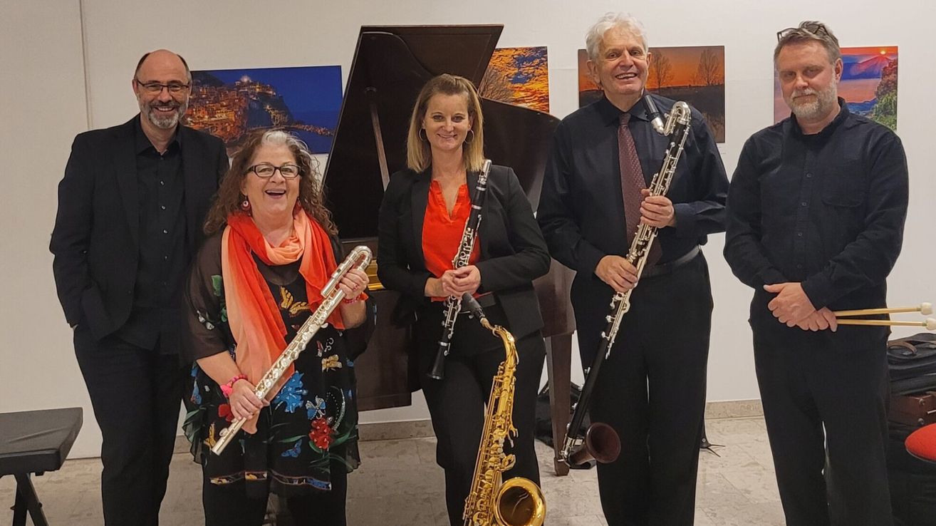 Three musicians stand next to a piano, smiling for a photo. A woman holds a flute, another a saxophone, and a man a clarinet. They are in a room with a gallery-like background.