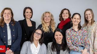 A group of seven women, dressed in various attire, smiling for a photograph against a plain background.