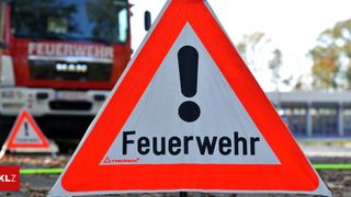 A white and red sign with the word 'Feuerwehr' and an exclamation mark is shown. A red fire truck is parked in the background.