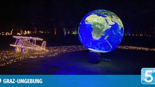 A large illuminated globe is displayed at night, with a trail of lights and a sleigh nearby. Trees and distant buildings are visible.