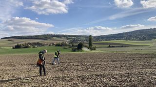 Two individuals in high-visibility vests are standing in a vast field with a road visible in the distance under a clear blue sky.