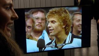A television screen displays a man named Gernot Jurtin, with a microphone in front of him, surrounded by others.
