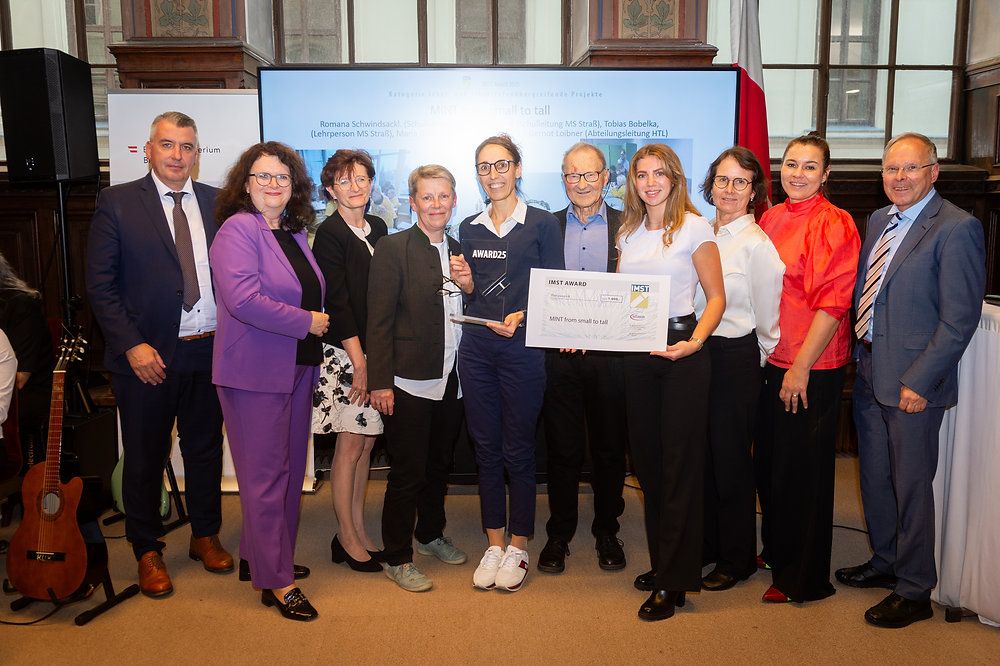 A group of individuals stands in a formal setting, likely at an award ceremony. They hold a certificate with the IMST Award logo. The woman in the center wears glasses and holds the certificate.