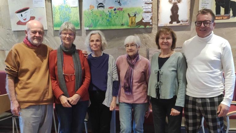 Vier Frauen stehen vor einer Wand mit Gemälden. Sie tragen Winterkleidung und Brillen. Die Frau auf der linken Seite trägt einen roten Pullover.