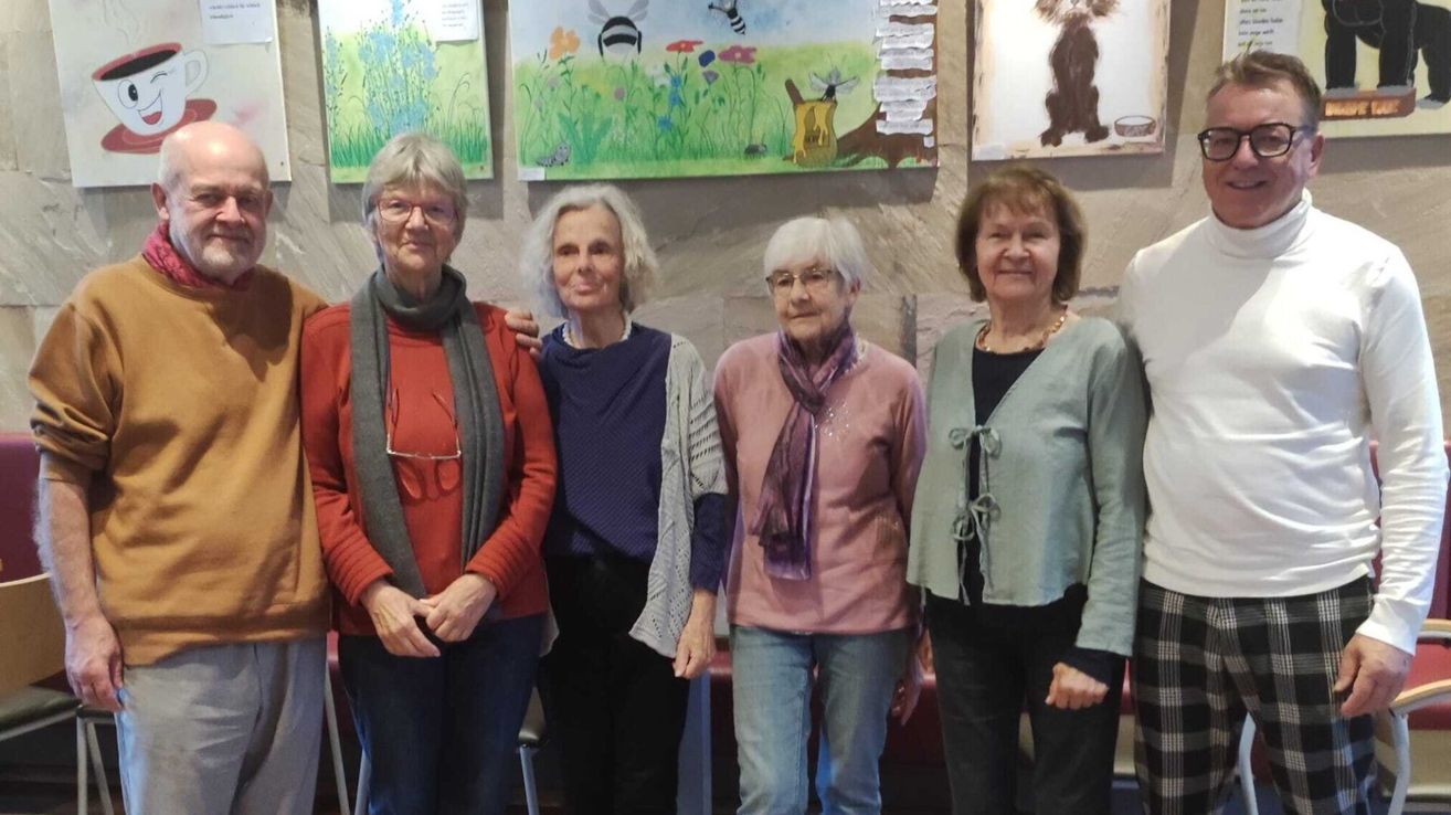 Vier Frauen stehen vor einer Wand mit Gemälden. Sie tragen Winterkleidung und Brillen. Die Frau auf der linken Seite trägt einen roten Pullover.