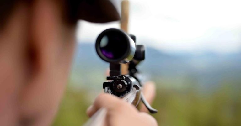 Eine Person zielt mit einem Gewehr und einem Zielfernrohr, hält es mit einer Hand, während die andere das Zielfernrohr einstellt. Der Hintergrund ist verschwommen mit einem Grasbereich und entfernten Bergen.