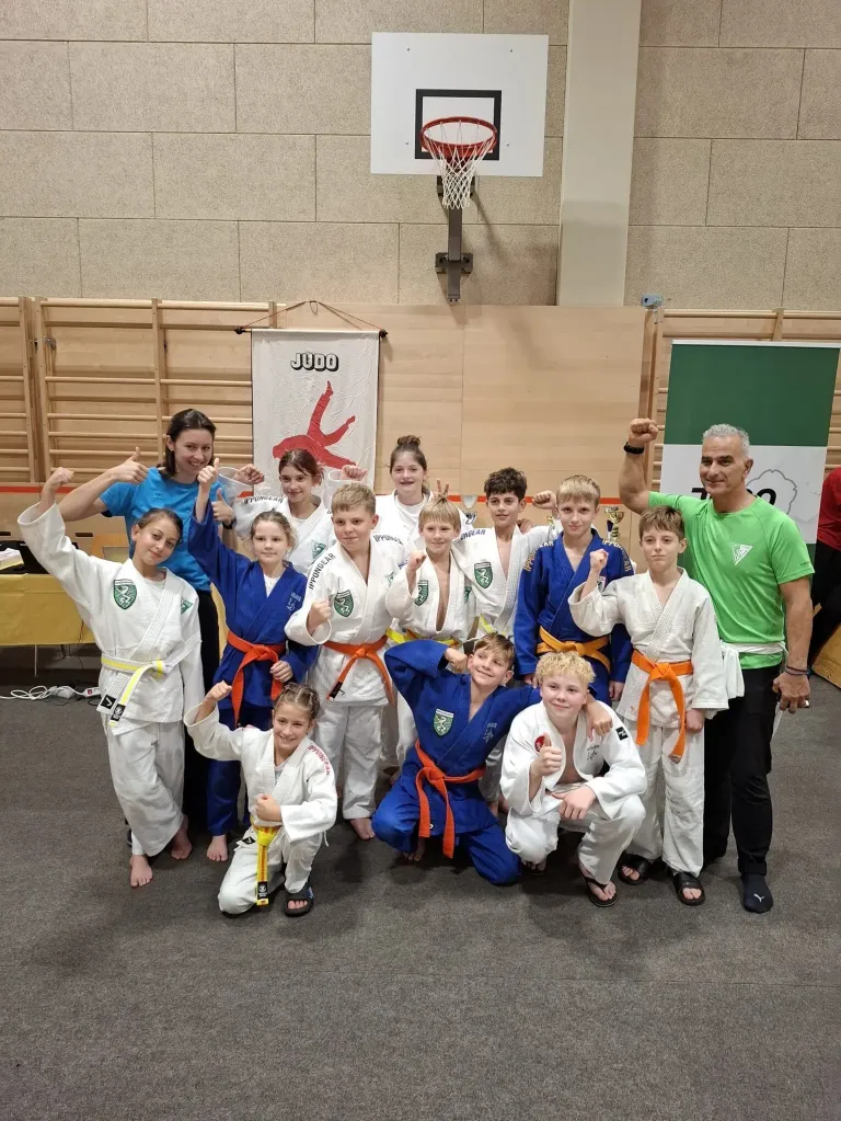 Eine Gruppe junger Judokas in blauen und weißen Uniformen posiert für ein Foto mit einem Mann in einem grünen Shirt. Sie stehen in einer Turnhalle mit hölzernen Wänden und einem Basketballkorb im Hintergrund.