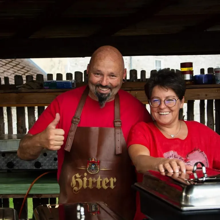 A man and a woman, both wearing red shirts, are standing in a kitchen. The man is giving a thumbs up and wearing an apron with the word 'Sirter' on it. The woman is smiling and holding a silver container.