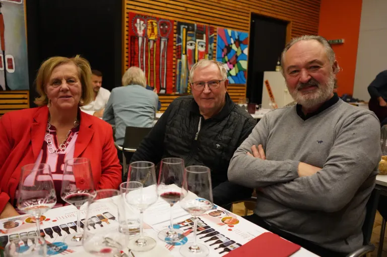 Drei Personen, zwei Männer und eine Frau, sitzen um einen Tisch mit Weingläsern und einer Speisekarte. Der Hintergrund zeigt farbenfrohe abstrakte Kunst.