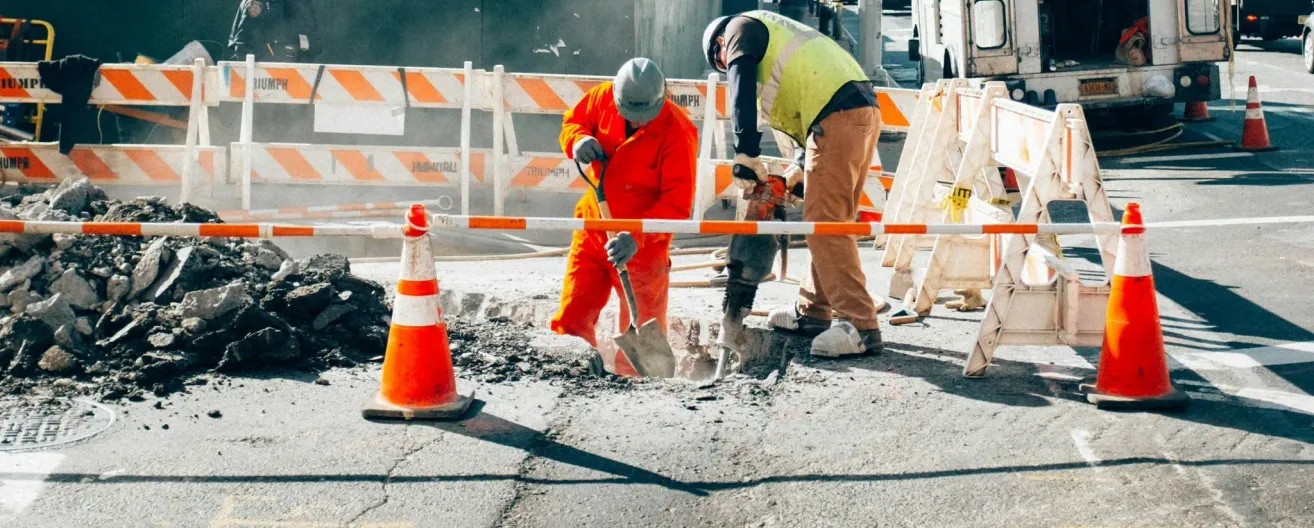 Two construction workers are working on a road. One is digging a hole with a shovel, and the other is using a power drill. A traffic cone is nearby.
