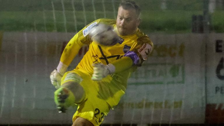 A soccer player in a yellow uniform is diving to block a ball in a nighttime match.