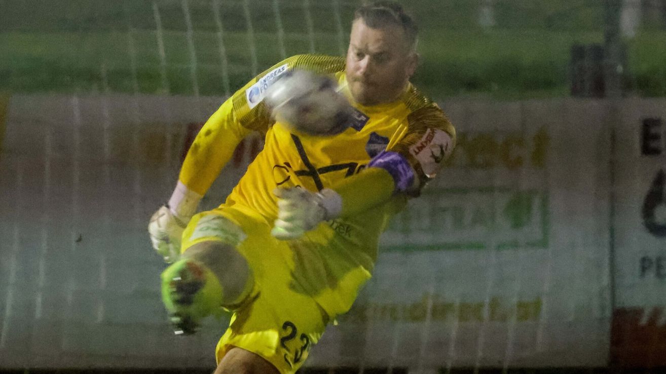 A soccer player in a yellow uniform is diving to block a ball in a nighttime match.
