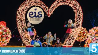Children pose for a photo in front of a heart-shaped structure adorned with lights and the logo 'eis GREISSLER' at night.
