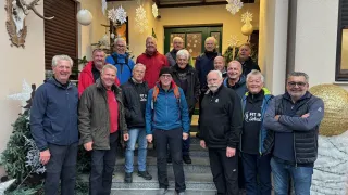 A group of older men wearing winter jackets and pants pose for a photo in front of a house decorated with Christmas ornaments and snowflakes.