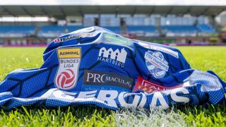 A blue and white sports jersey with multiple sponsor logos lies on a grassy field in front of an empty stadium.