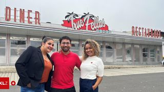 Three people are standing in front of a diner with a red roof and sign that reads True Fellas Diner. Two women and one man are smiling and posing for a photo.