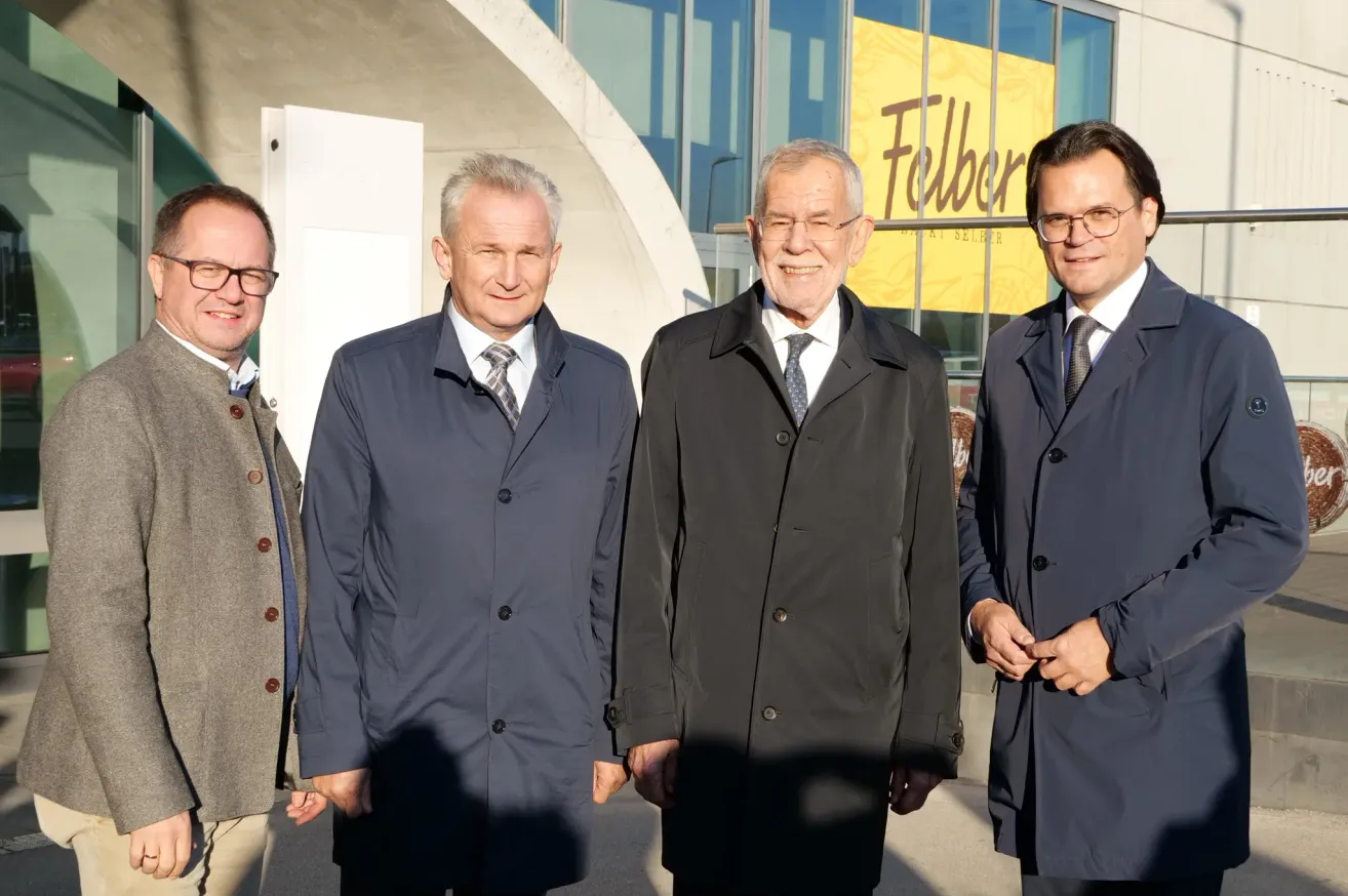 Four men in formal attire stand together outside a building with a yellow banner reading 'Felber'. They are smiling and appear to be posing for a photograph.