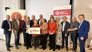 A group of people stands in a room, smiling for a photo. They hold a sign that reads 'pensionisten verband'. Behind them, a banner with the word 'Melk' is visible.