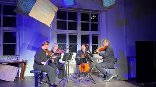Four musicians are performing on stage with violins and a cello, illuminated by purple lights. Two women sit with violins, while two men play the cello. The stage is decorated with hanging papers and a white backdrop.